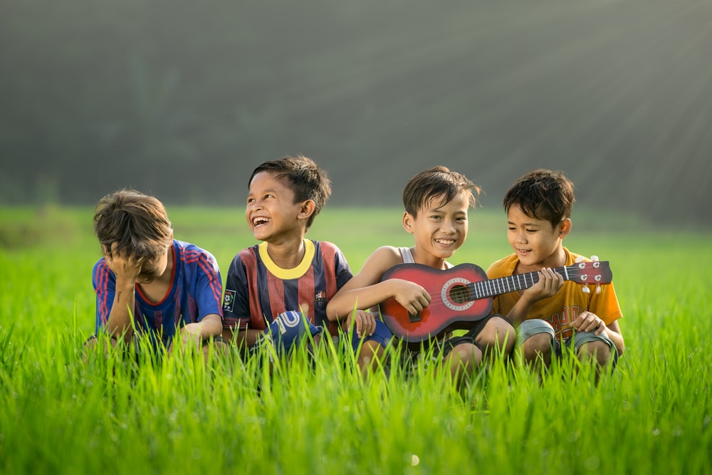 Four boys sitting on the grass, playing the guitar, and laughing during daytime